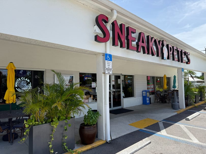 Exterior of Sneaky Pete's restaurant with outdoor seating, planters, and a parking lot.